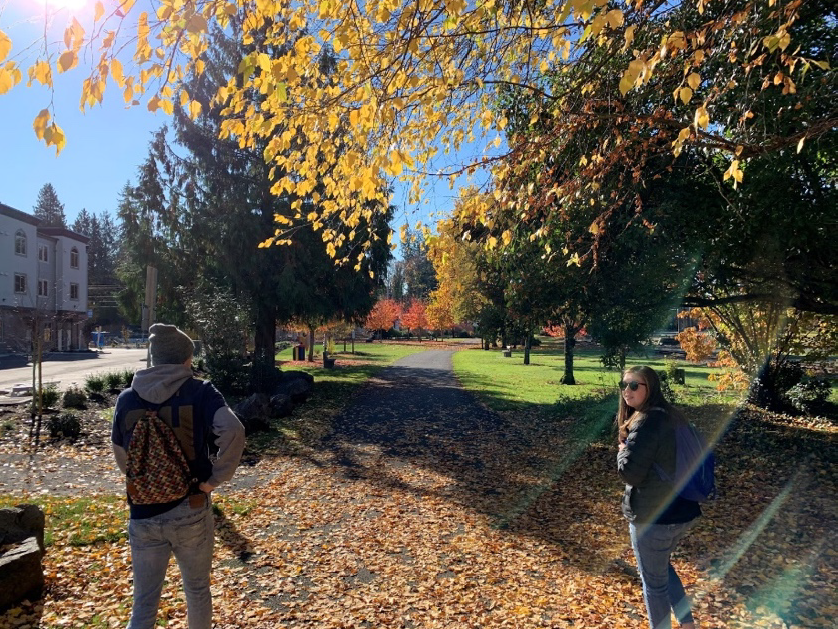 Two students standing on fallen leaves on Centennial Trail under an autumn-colored tree