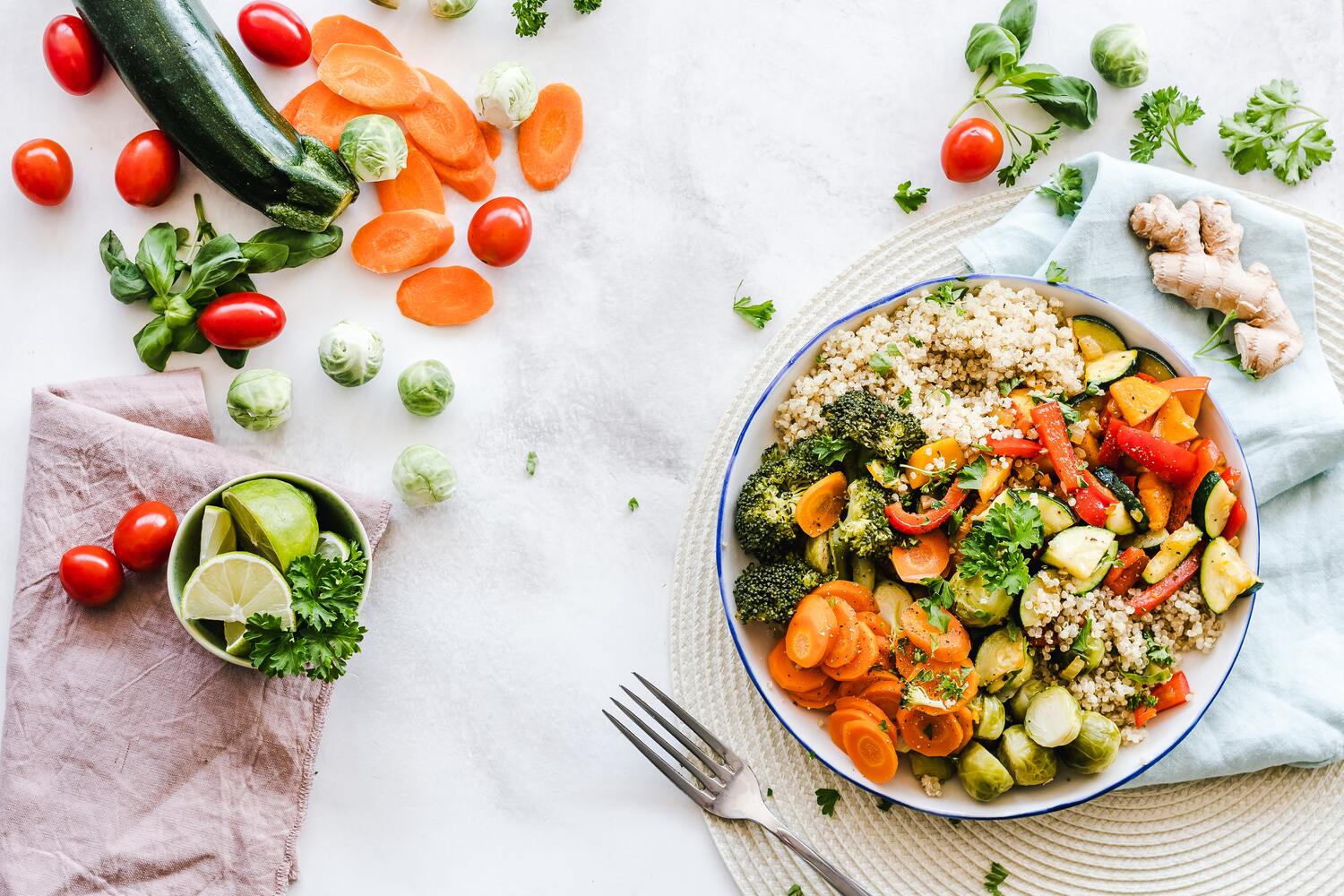 An image showing a bowl of quinoa and cooked vegetables, garnished with herbs, on a table surrounded by raw zucchini, carrots, brussels sprouts, cherry tomatoes, lime wedges, and a fork beside the bowl.