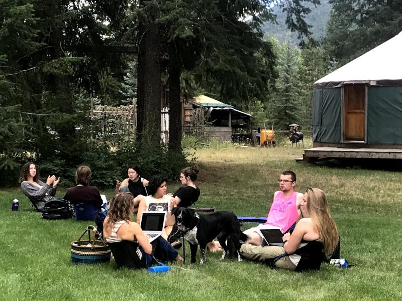 Students sit in a field, working on a group project on laptops outside a yurt