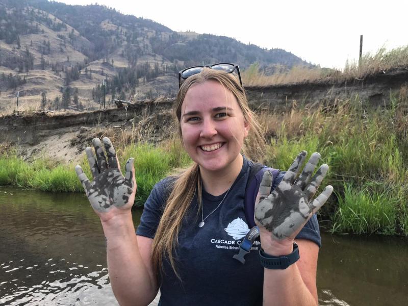 Student Katie Boon holds up mud-caked hands in front of a river